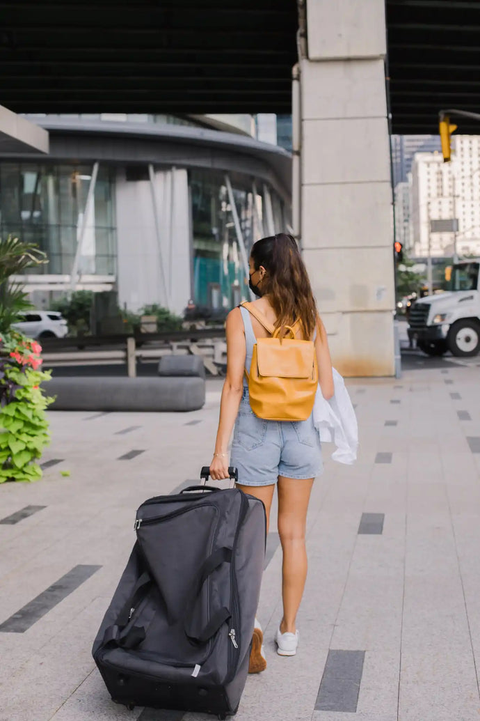 Une personne tirant une valise à roulettes noire tout en portant un sac à dos jaune.